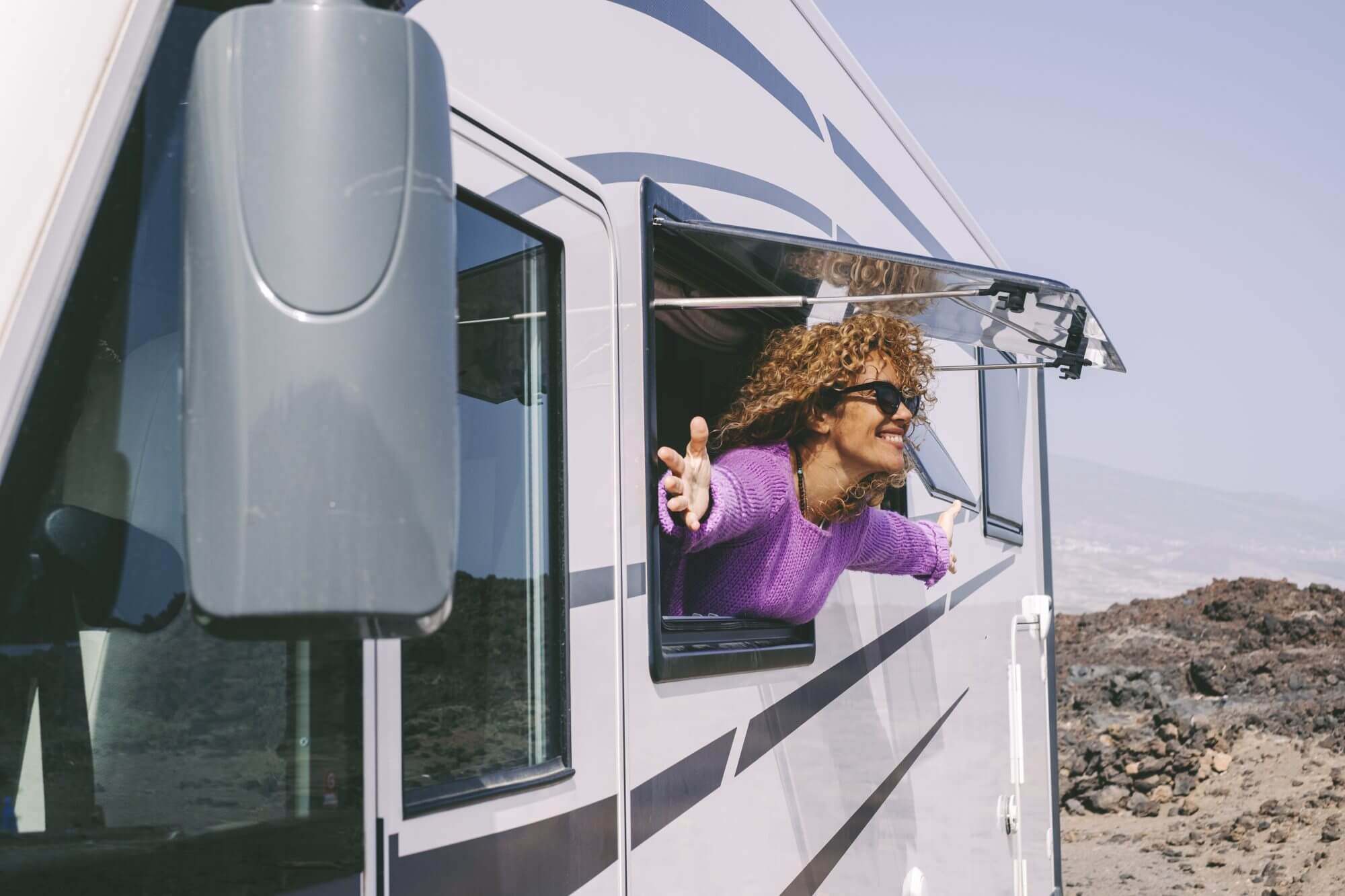 Overjoyed and excited adult pretty woman admire outside the camper van window and outstretching arms with big smile. Concept of tourist people and tourism with rented vehicle. Nature campsite