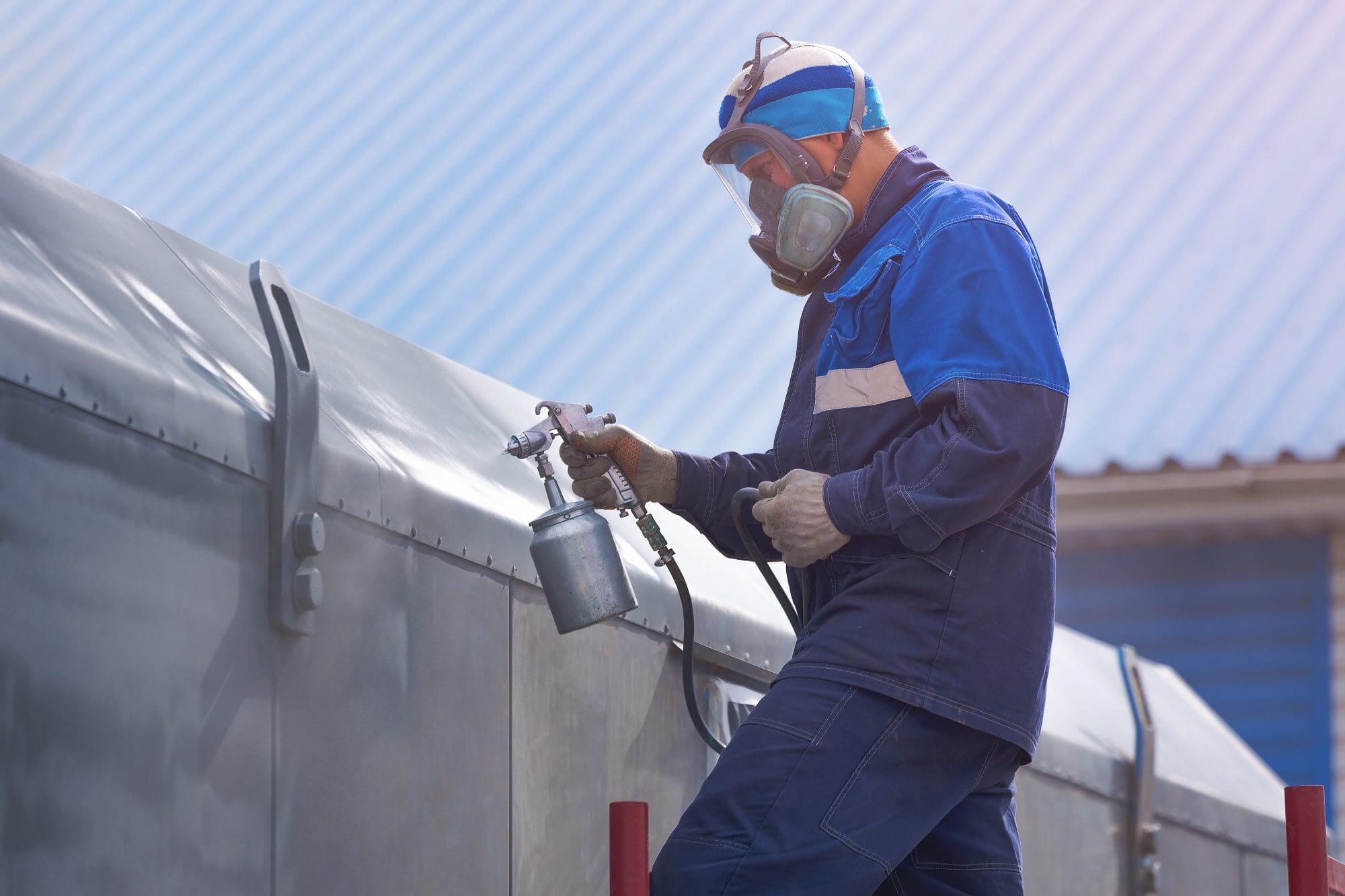 Industrial work. Priming of metal products from the compressor gun. A worker in overalls and a respirator stands on the stairs and paints the body of a truck trailer or a metal car. Work at height