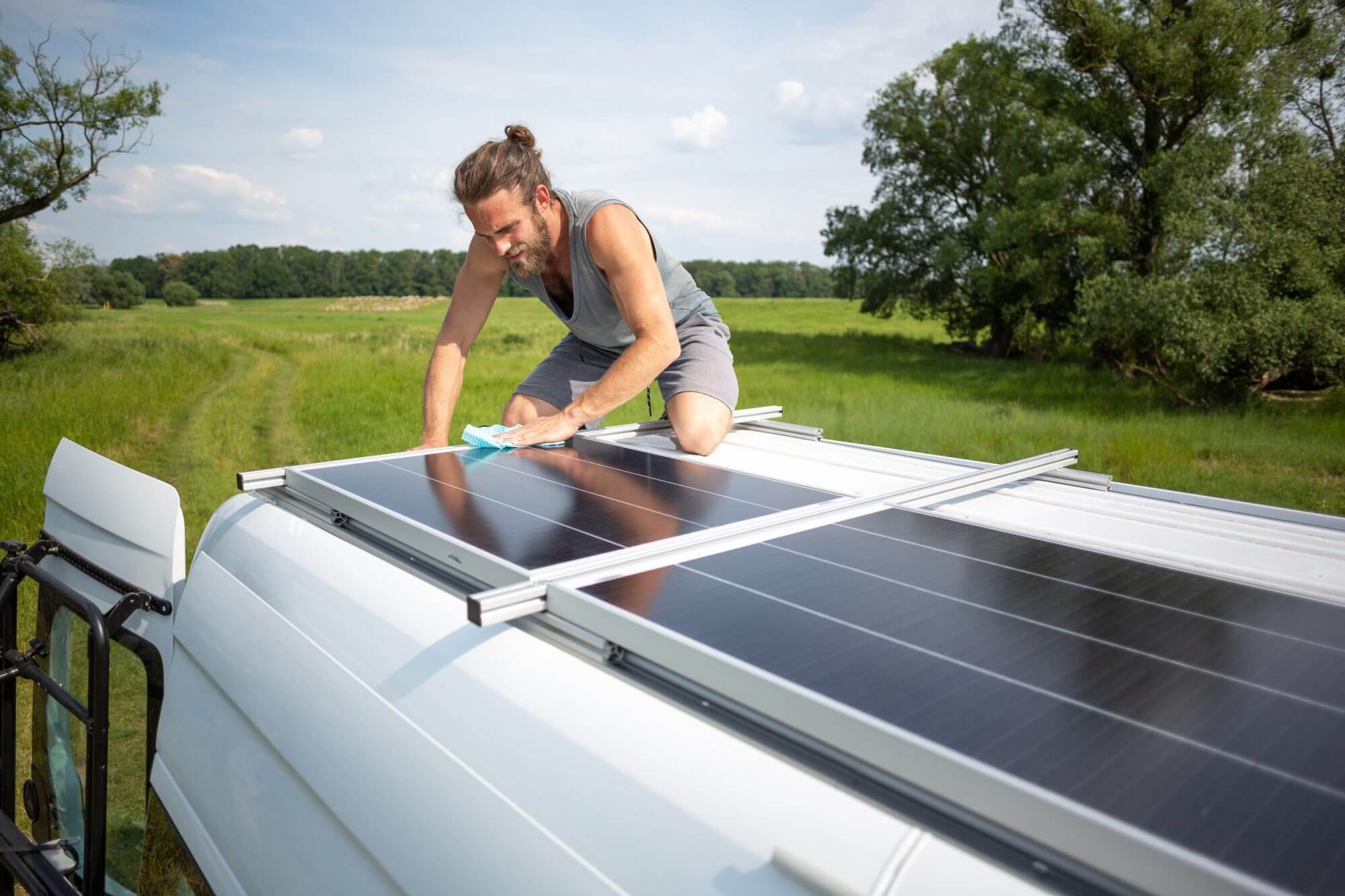 Young man on top of the roof of a camper van with solar panels. He is using a cloth to wipe clean the panels. The sun is shining and a green meadow with trees is visible in the background.