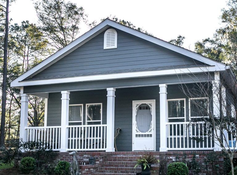 Small blue gray mobile home with a front and side porch , white railing out in the country on land shared by a larger siding house.