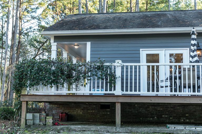 Side view of small blue gray mobile home with a front and side porch with white railing, out in the country on land shared by a larger siding house.