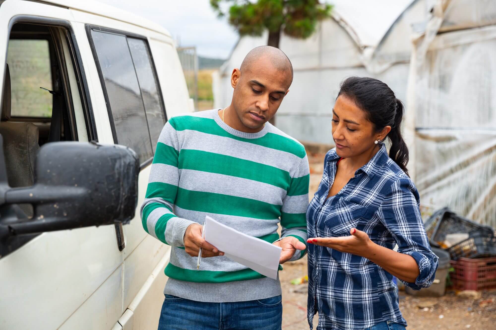 Hispanic couple of farmers discussing some papers while standing near car on backyard of farmstead