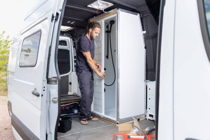 A van with an open door. A young man is standing inside installing a small shower room. Work in progress converting a van into a camper van.