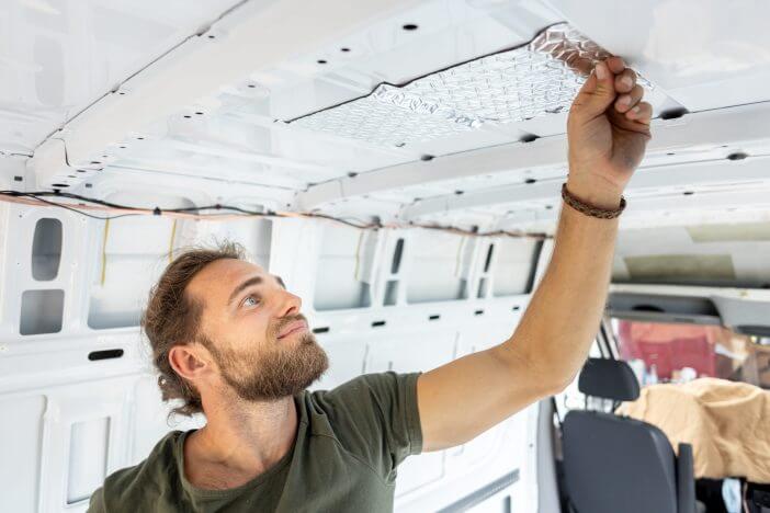 Young man standing inside a van. He is attaching insulation material onto the roof. Camper van conversion project.