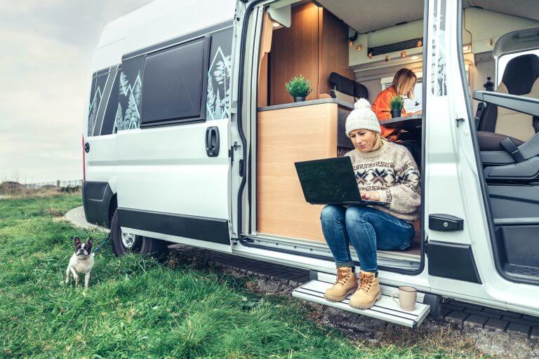 Woman teleworking sitting in the door of a camper van while her partner works inside