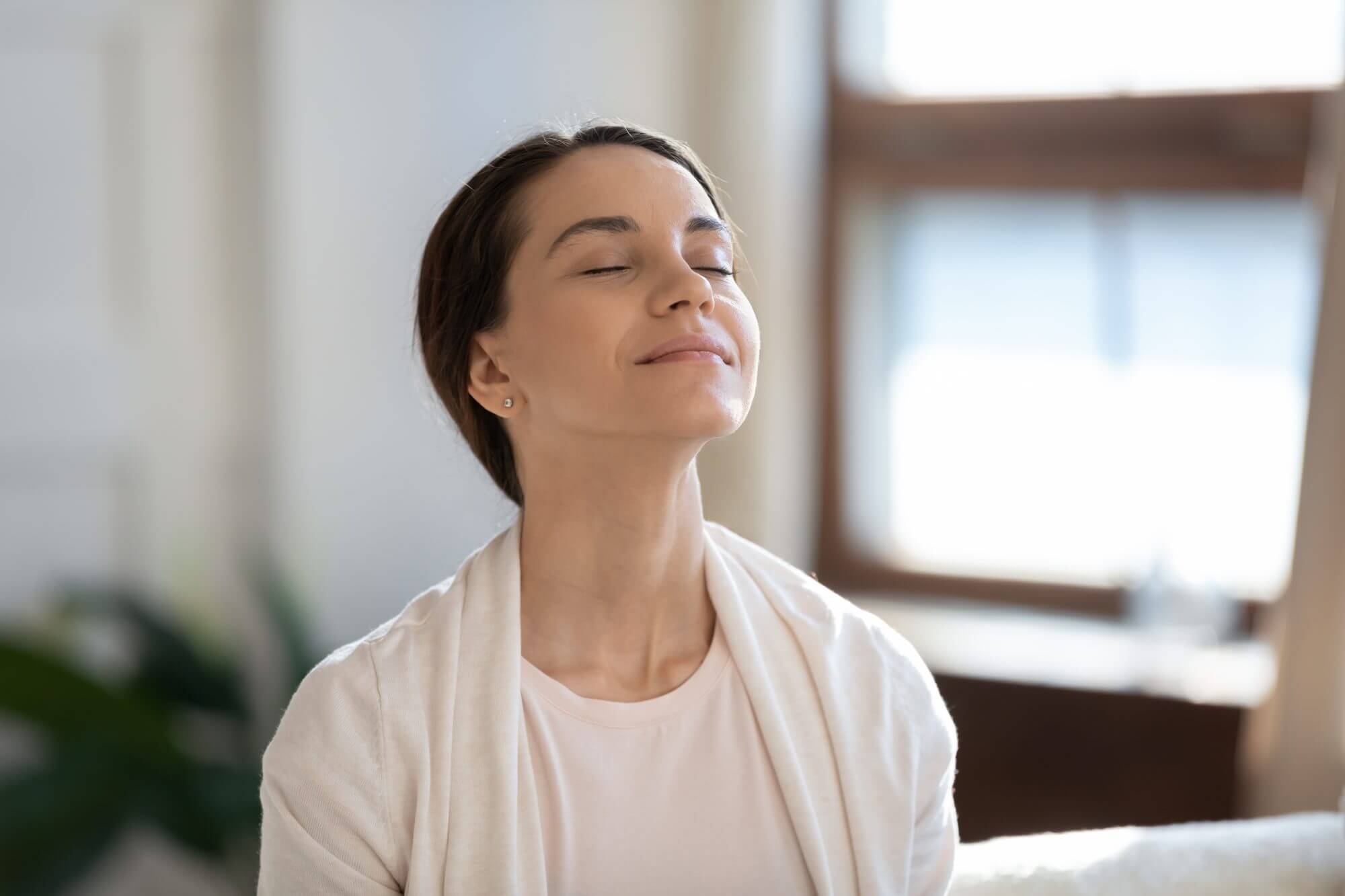 Calm leisure. Content young woman spending time at home alone, sitting on sofa with eyes closed in delight, breathing fresh cool air because of working conditioner, giving face to sunlight from window