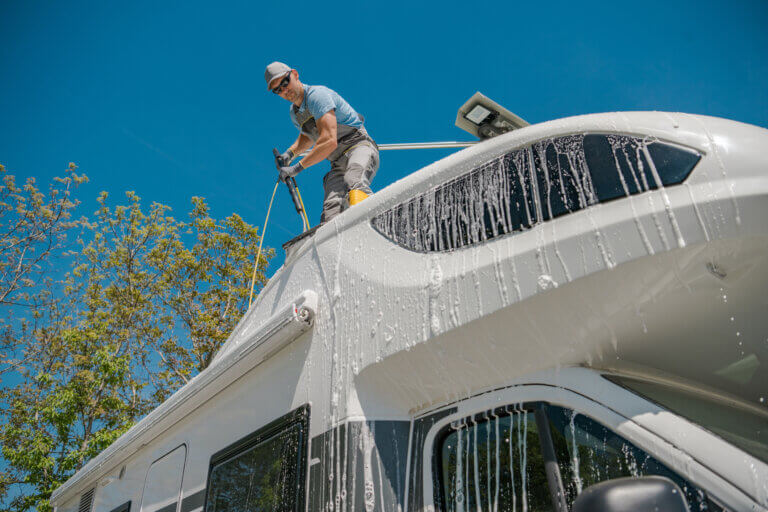 Caucasian Man Washing His White Motorhome While Standing on the Top of Its Roof. Camper Van Owner Taking Care of His Vehicle After a Long Trip.