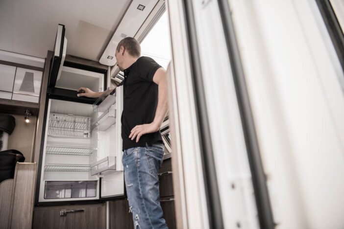 Broken RV Camper Travel Trailer Refrigerator. Caucasian Technician Trying to Fix the Appliance Inside Modern Recreational Vehicle.
